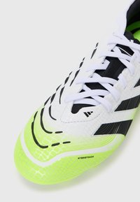 White athletic shoe with black and neon green accents. Features textured synthetic material, lace-up design, and ribbed sole with "HYBRIDTOUCH" detail.