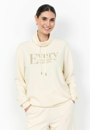 Uśmiechnięta kobieta o ciemnych włosach nosi kremową bluzę i pasujące spodnie, ręce w kieszeniach, stoi na tle jednolitego białego tła.