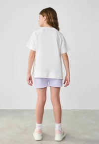 Camiseta blanca de algodón con mangas cortas, combinada con pantalones cortos de color púrpura claro. Costuras visibles y un diseño de corte relajado. Fondo neutro.