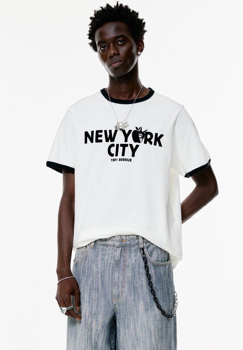 Hvid bomulds t-shirt med sort krave og manchetter, prydet med en markant "NEW YORK CITY" grafik og kædeaccent på denimbukser.