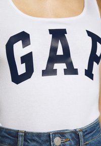White ribbed tank top featuring large dark blue letters spelling "GAP," paired with blue denim shorts.