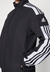 Black athletic jacket with high collar, front zipper, and contrasting white stripes on sleeves. Features elastic cuffs and side pockets.