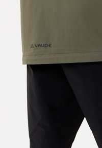 Olivgrünes kurzärmliges Hemd aus leichtem Stoff, mit geradem Saum und einem Logo am unteren linken Rand. Schwarze Hosen sind teilweise sichtbar.