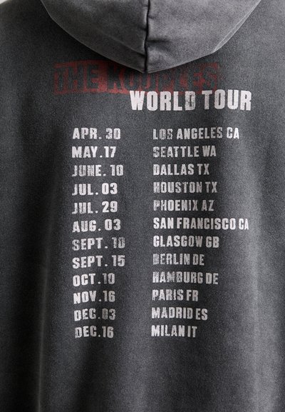 Ciemnoszara bluza z kapturem z napisem "THE KOURLES WORLD TOUR" w wyraźnym czerwonym i białym kolorze. Zawiera listę miast i dat w przejrzystym, uporządkowanym formacie.