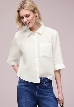 Lyshåret kvinde iført en løs hvid skjorte med trekvartærmer og blå denimjeans, der kigger nedad og smiler.