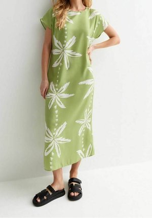 Kvinde i en lysegrøn midikjole med store hvide blomsterprint, poserer med den ene hånd på hoften og iført sorte platformsandaler.