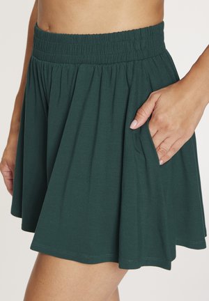 Frau trägt einen dunkelgrünen Rock mit elastischem Bund, knielang, mit der Hand in einer Seitentasche vor hellem Hintergrund.