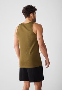 Olivgrünes Tanktop mit geripptem Muster, ärmellosem Design und rundem Ausschnitt, kombiniert mit schwarzen Shorts. Modell von hinten gezeigt.