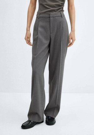 Person iført grå bukser med høj talje og vide ben med læg, mørk top og sorte loafers, stående mod en ensfarvet lys baggrund.