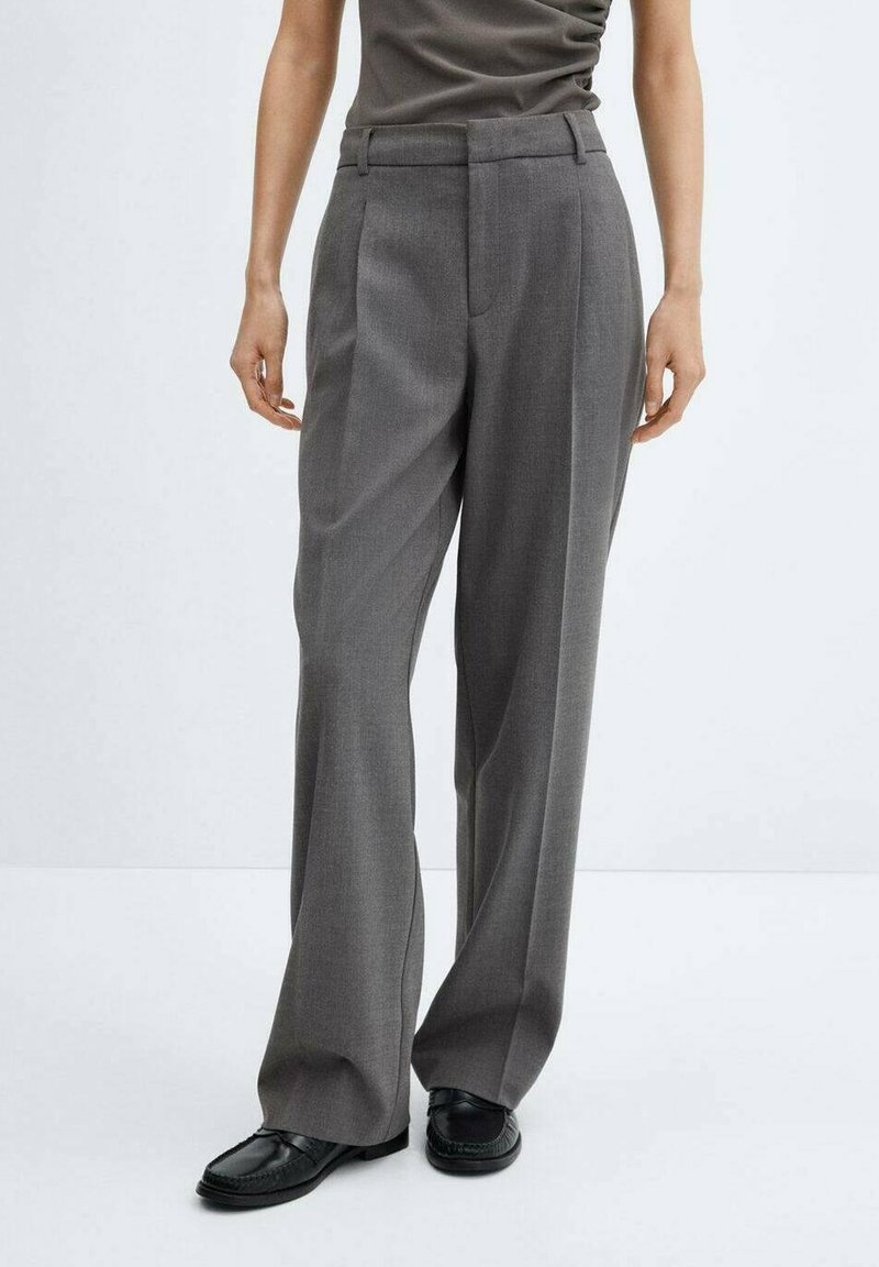 Personne portant un pantalon gris taille haute à jambes larges avec des plis, un haut foncé et des mocassins noirs, debout devant un fond clair uni.