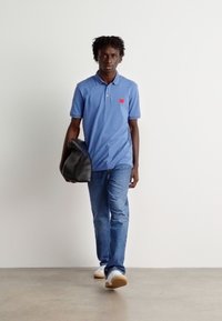 Blå poloskjorte med en rød logo, denimjeans og hvite joggesko. Holder en svart skinnveske, står mot en ensfarget bakgrunn.