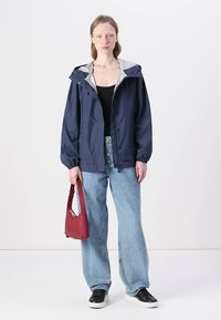 Giacca impermeabile blu navy con cappuccio grigio, vestibilità comoda e polsini elasticizzati, abbinata a jeans a gamba larga blu chiaro e una borsa rossa.