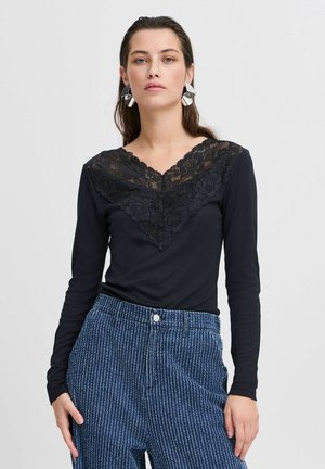 Schwarzes Langarmoberteil mit einem Spitzen-V-Ausschnitt, das mit floralen Mustern verziert ist. Kombiniert mit blau gestreiften weit geschnittenen Hosen.