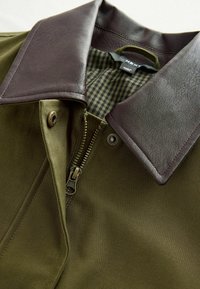 Olivgrüne Jacke mit einem dunkelbraunen Kunstlederkragen, vorderem Reißverschluss und kariertem Innenfutter. Akzentknopf in der Nähe des Reißverschlusses.