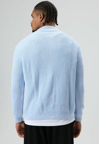 Homme aux cheveux tressés portant un pull en maille bleu clair, un sous-vêtement blanc et un pantalon noir, vu de dos sur un fond gris uni.