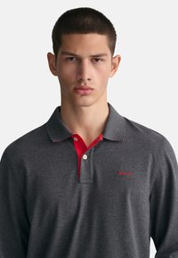 Graues Langarm-Poloshirt mit rotem Kragen. Mit kleinem rotem Logo auf der linken Brust und subtilen Kontrastnähten.