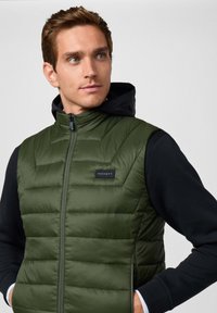 Zielona kamizelka ocieplana z zamkiem z przodu, czarnym kapturem i naszywką z logo. Posiada pikowaną strukturę i boczne kieszenie, noszona na czarnej bluzie z długim rękawem.