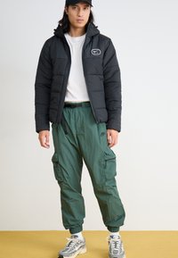 Μαύρο μπουφάν puffer με κουκούλα και λογότυπο, σε συνδυασμό με χαλαρές πράσινες cargo παντελόνες και γκρι αθλητικά παπούτσια. Το σύνολο έχει μια χαλαρή, λειτουργική σχεδίαση.