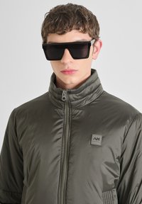 Giacca puffer verde scuro con zip, colletto alto e patch con logo; chi la indossa ha occhiali da sole neri oversize e capelli ricci ben curati.