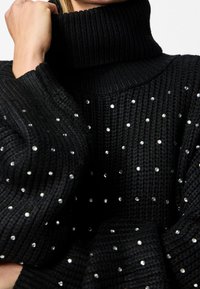 Svart stickad tröja med hög ribbad krage och spridda rhinestone-utsmyckningar som skapar en texturerad yta.