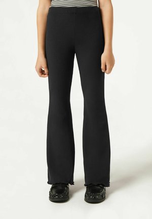 Pantalons évasés côtelés noirs avec une taille haute, dotés d'un tissu texturé et d'une finition ourlet brut, portés avec des chaussures noires.