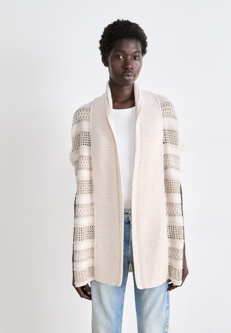 Hellbeige, offenes Strickjacke mit gestreiftem Muster, weicher Textur und weiten Ärmeln. Getragen über einem weißen Oberteil und kombiniert mit hellblauen Jeans.