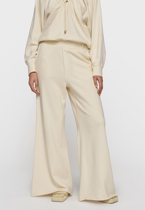 Kobieta ubrana w kremowe, szerokie spodnie z wysokim stanem i luźnym krojem, zestawione z kremową bluzką i płaskimi butami.