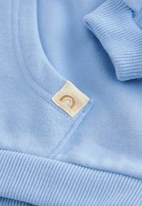 Lichtblauwe katoenen kleding met ribbelmanchetten, voorzien van een gestikt label met een beige achtergrond en een bruin boogontwerp.