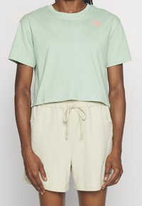 Světlé zelené krátké tričko s krátkými rukávy a růžovým logem na levé straně hrudi, doplněné béžovými šortkami s tkanicí. Hladká textura látky.