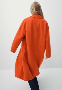Lungo cappotto arancione con colletto a revers, vestibilità ampia e spalle basse. Tessuto morbido, design fino al ginocchio e due tasche laterali.