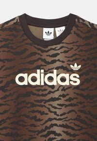 Barna terepmintás rövid ujjú póló kerek nyakkivágással, fehér "adidas" logóval és egy kis fehér levél logóval.