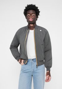 Γκρι καπιτονέ bomber jacket με ριχτές μανσέτες, κίτρινες και λευκές λεπτομέρειες στο φερμουάρ και ένα μικρό λογότυπο στον αριστερό ώμο. Φοριέται πάνω από μια λευκή μπλούza.
