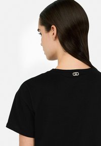 Schwarzes Baumwoll-T-Shirt mit Rundhalsausschnitt und kurzen Ärmeln, das ein kleines goldenes Logodetail auf dem Rücken nahe dem Ausschnitt hat.