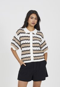 Sweter robiony na drutach z kołnierzykiem, w naprzemienne wzory w odcieniach beżu, bieli i czerni. Z przodu zapięcie na guziki i krótkie rękawy.