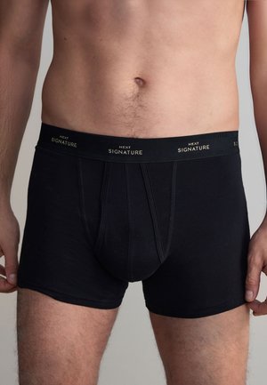 Mies vartalo mustat bokserit joissa vyötäröllä teksti "Next Signature", seisoo yksiväristä harmaata taustaa vasten.