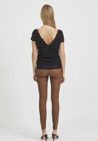 Schwarzes Spitzenoberteil mit einem gewellten Halsauschnitt, kombiniert mit braunen, schmalen Hosen und schwarzen High Heels. Die Haare sind glatt frisiert und fallen den Rücken hinunter.