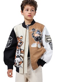 En ung dreng iført en multicolor basketballjakke med en tegneserietiger og en fugl, der spiller basketball, beige bukser, står foran en hvid baggrund.