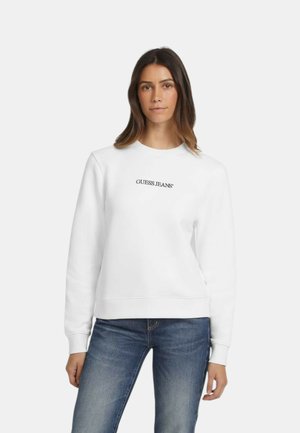 Donna con capelli castani lisci che indossa una felpa bianca con la scritta "GUESS JEANS" e jeans blu, in piedi contro uno sfondo semplice.