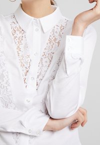Weißes Hemd mit Knopfverschluss, das mit Spitzenpaneelen auf der Vorderseite, einem klassischen Kragen und langen Ärmeln mit Knopfmanschetten ausgestattet ist. Glatte Stofftextur.