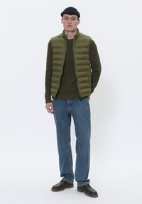 Maslinasto zeleni pufer prsluk preko teksturirane tamnozelene veste, uparen s svijetloplavim trapericama i smešnim cipelama, stoji naslonjen na bijelu pozadinu.