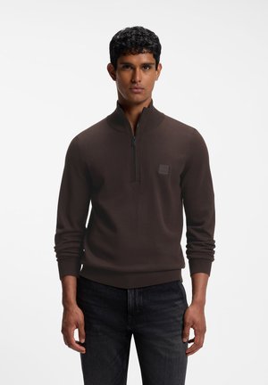 Homme portant un pull marron à manches longues avec demi-fermeture éclair et un jean noir, debout les bras détendus devant un fond blanc.