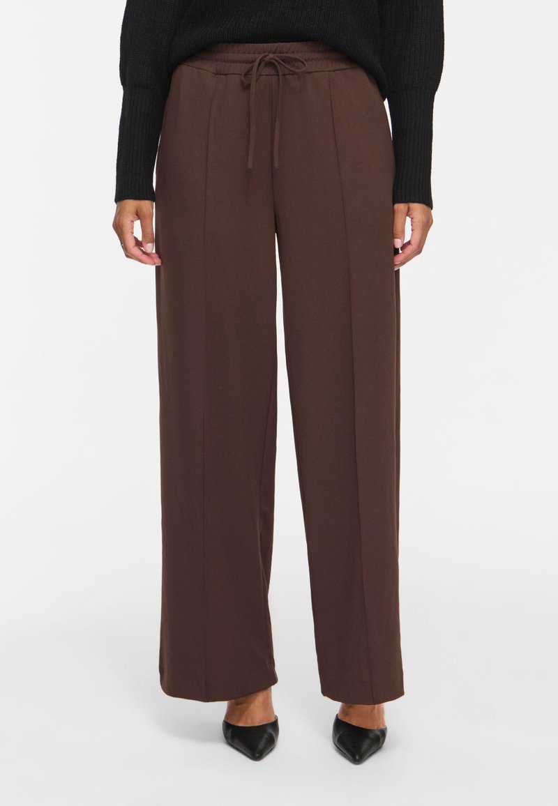 Femme portant un pantalon large marron foncé avec une taille à cordon et des chaussures noires pointues, debout devant un fond blanc uni.