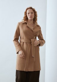 Brun ullfrakk med belte i midjen, spiss krage og skrå glidelås. Glatt tekstur, skreddersydd design og lange ermer.