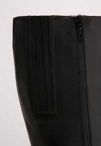 Lähikuvassa sileä musta nahkasaapas, jossa on pystysuorat venykkeet ja musta vetoketju sivussa.