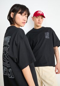 Black cotton t-shirts with oversized fit; one features a detailed graphic print on the back, while the other has a small front emblem.
