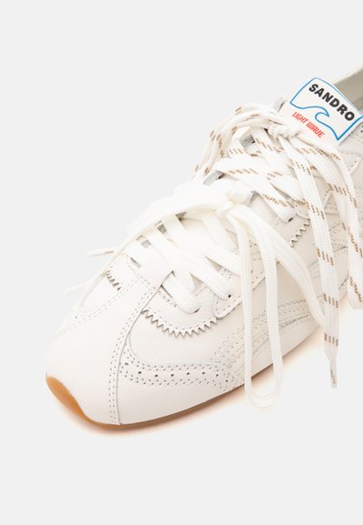 Gros plan d'une basket en cuir blanc avec une couture en zigzag, des lacets doubles blancs et beiges, et une étiquette "SANDRO Light Wave" sur la languette.