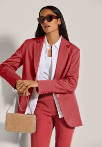 Roter Blazer und passende Hose mit einem weißen Hemd darunter. Die Accessoires umfassen eine beigefarbene Handtasche mit einer Kettenriemen und übergroße Sonnenbrillen.
