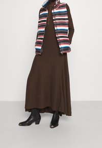 Ruskea pitkähihaista mekkoa on yhdistetty moniväriseen raidalliseen pufferi-vestiin ja mustiin korkokenkiin, joissa on hopeiset kärjet.