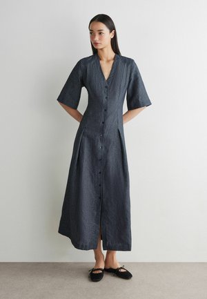 Kvinde stående med hænderne bag ryggen iført en mørkegrå knap foran midi-kjole med albuelange ærmer og sorte flade sko.