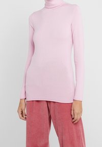 En person som bär en figursydd ljusrosa polotröja och lösa rosa kostymbyxor, som står mot en enfärgad ljus bakgrund.